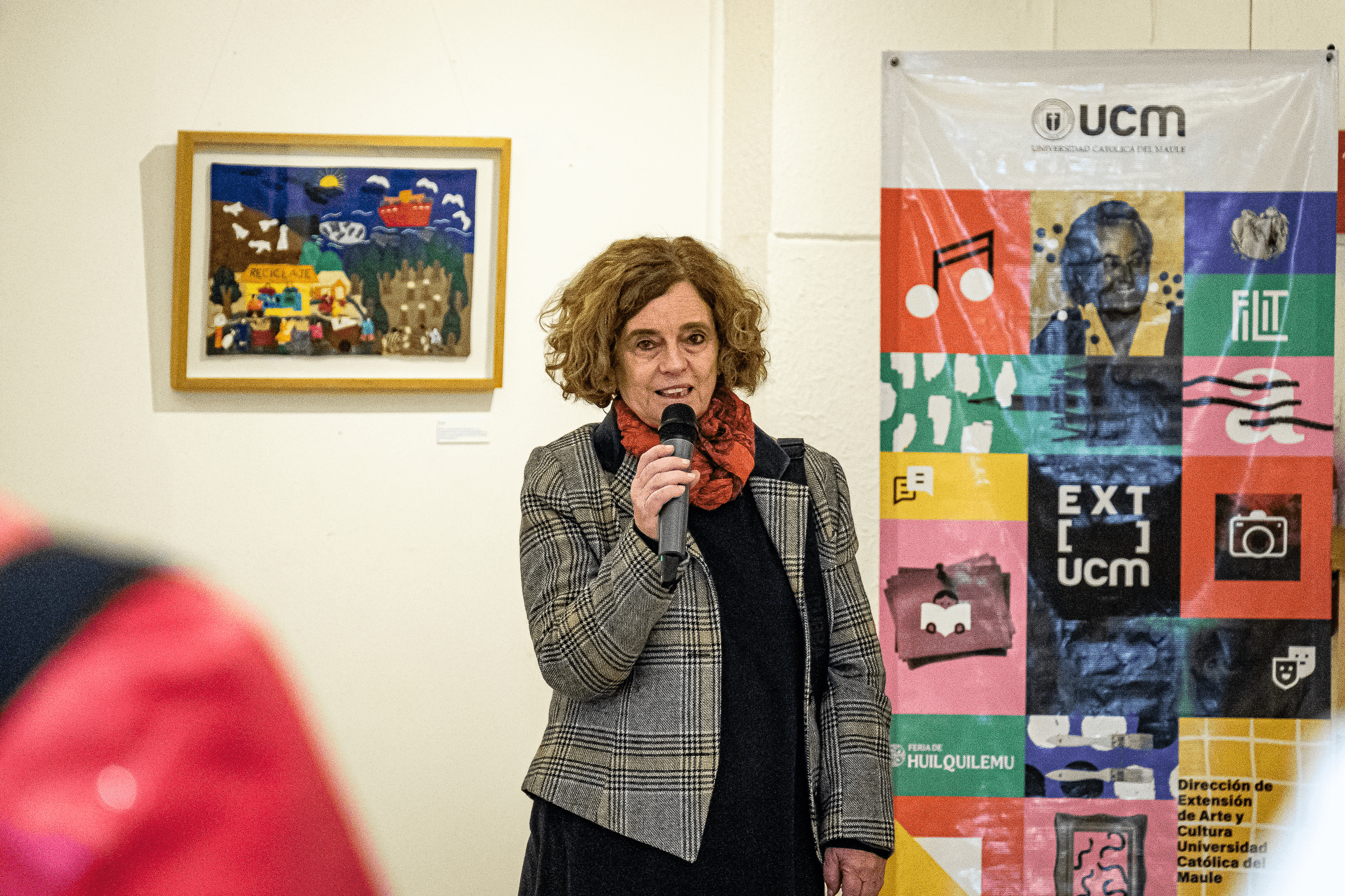 Una mujer está hablando con un micrófono en una exposición artística con obras al fondo.