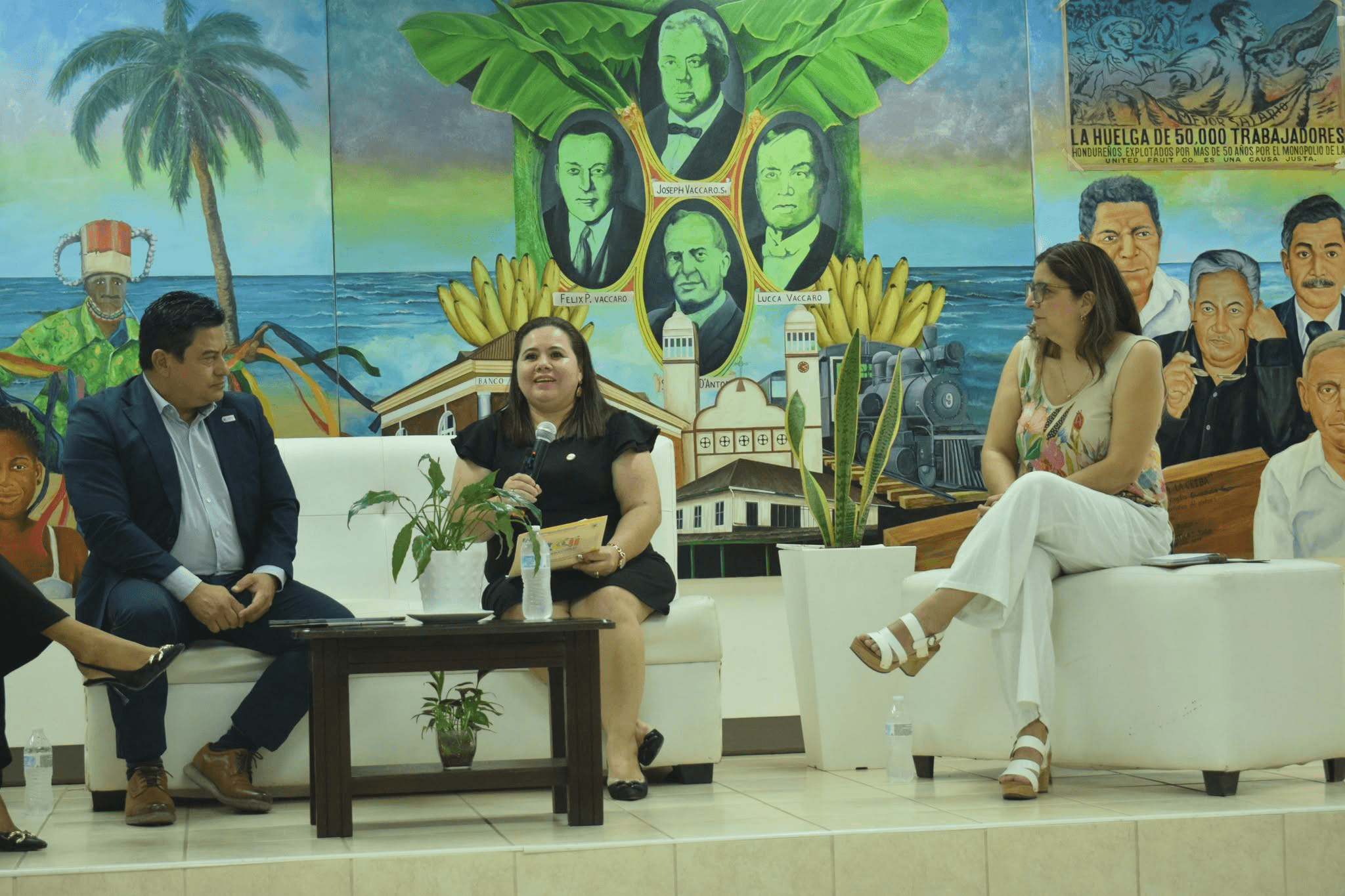 Un grupo de personas discute en un evento cultural con un mural colorido de fondo.