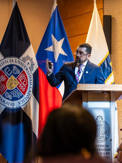 Un expositor presenta en una conferencia con banderas chilenas de fondo.