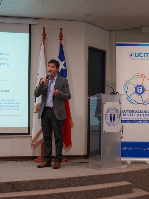 Un hombre presenta un informe sobre la autoevaluación institucional en una sala con banderas chilenas de fondo.