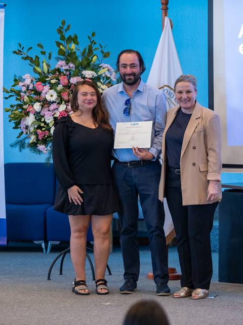 Una ceremonia de entrega de certificados donde un hombre sostiene su diploma entre dos mujeres.