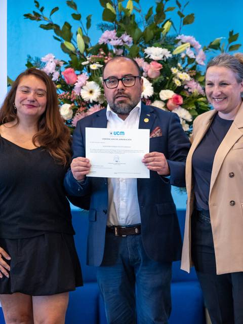 En la imagen se ve a un hombre sosteniendo un certificado junto a dos mujeres en un evento formal.
