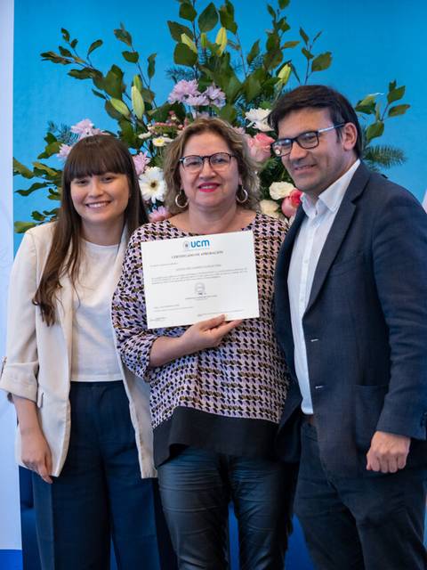 Una mujer recibe un diploma en una ceremonia con dos personas a su lado y un fondo decorado.