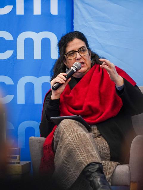 Una mujer con abrigo negro y chal rojo habla con entusiasmo en un panel dirigido por la universidad.