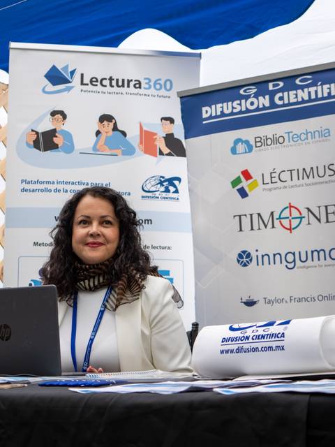 Una mujer sonriente está sentada en un stand de difusión científica en un evento.