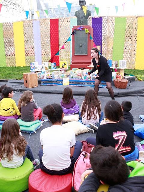 Una narradora cuenta historias a un grupo de niños sentados en cojines de colores en un ambiente festivo.