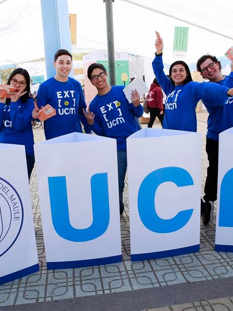 Un grupo de seis personas sonrientes sostiene folletos y está frente a grandes letras que representan la Universidad Católica del Maule en un ambiente festivo.