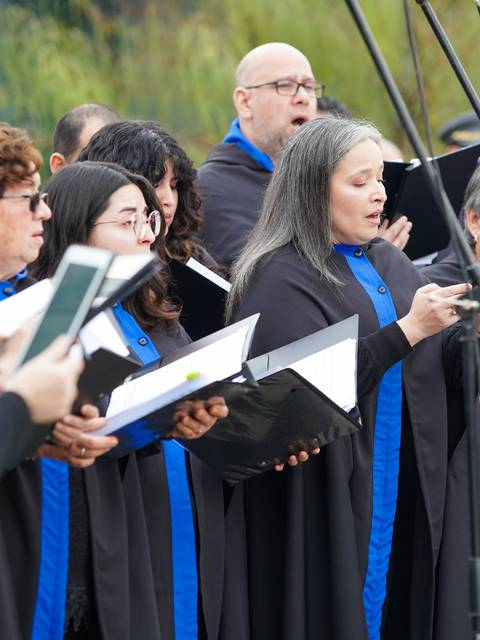 Un grupo de cantantes en un coro se presenta con capas negras y azules en un evento al aire libre.