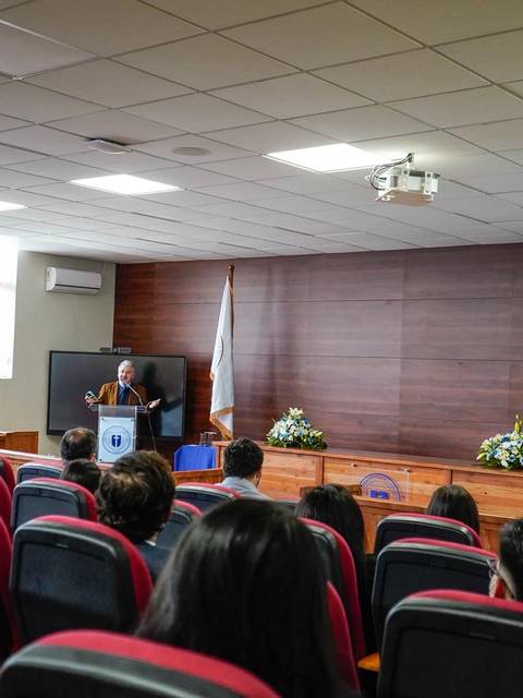 Un hombre habla en un auditorio con varias sillas ocupadas y una bandera chilena visible.