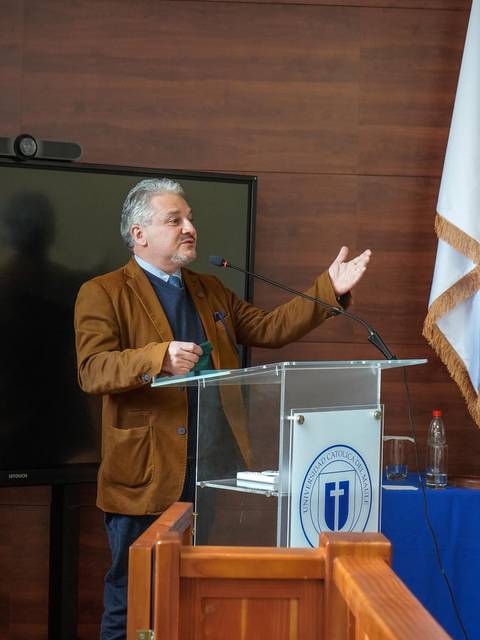 Un hombre está dando una charla en un podio frente a una pantalla y una bandera.