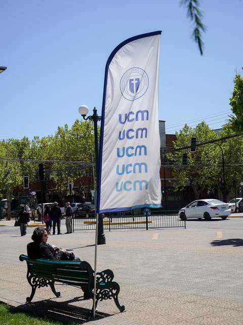 Una persona descansa en un banco junto a una bandera de la UCM en un día soleado.