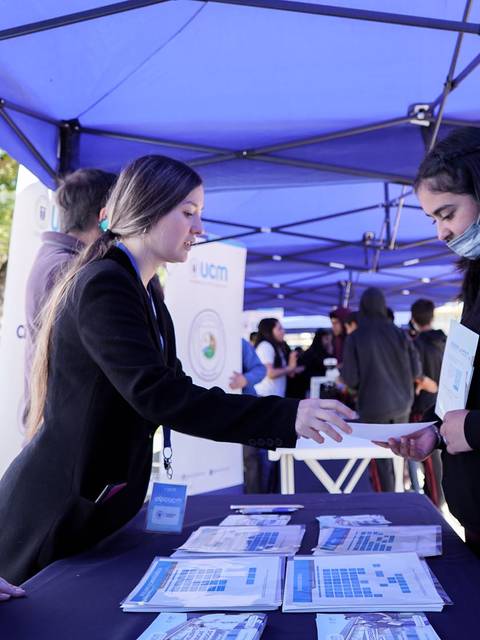 Un grupo de jóvenes interactuando en un evento bajo carpas, con material informativo sobre una universidad.