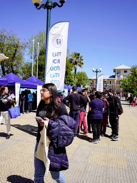 Una mujer camina entre un grupo de personas en un evento al aire libre con banderas y carpas.