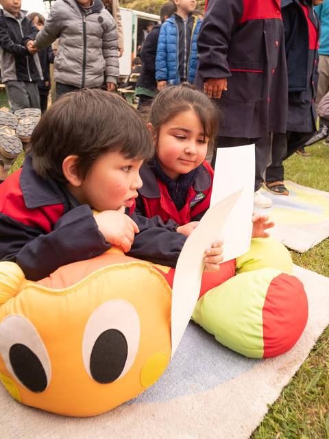 Dos niños se encuentran tumbados sobre el césped mirando un papel mientras están rodeados de otros niños y una atmósfera de juego.