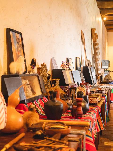 Un hombre explica frente a una mesa llena de artesanías y objetos culturales en un ambiente interior.