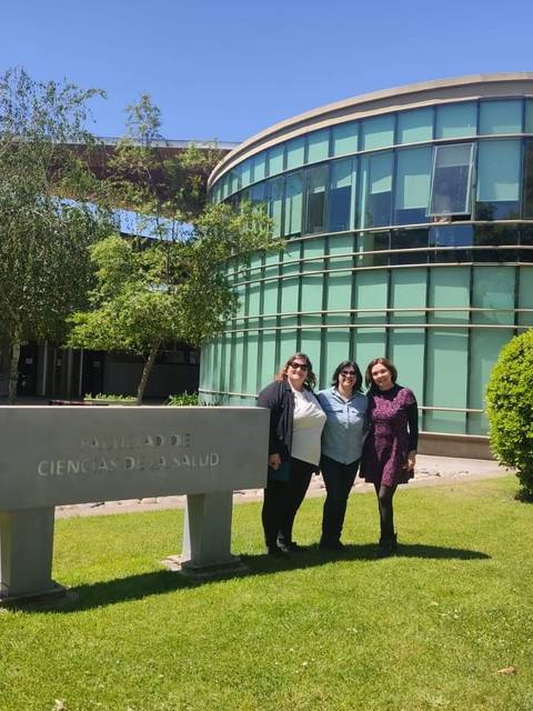 Tres mujeres posando frente a un edificio moderno de vidrio y hierba verde.