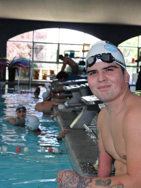 Un nadador sonriente con gorro y gafas de natación se sienta al borde de una piscina mientras otros niños nadan en el fondo.