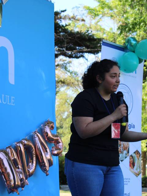 Una persona habla frente a un público en un evento al aire libre con globos y una decoración festiva.