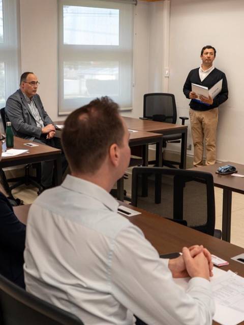 Una reunión de trabajo en una sala con varias personas sentadas y dos presentadores en pie.