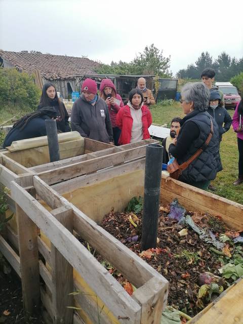 Un grupo de personas observa y escucha a una mujer que está explicando sobre la compostera en un entorno rural.