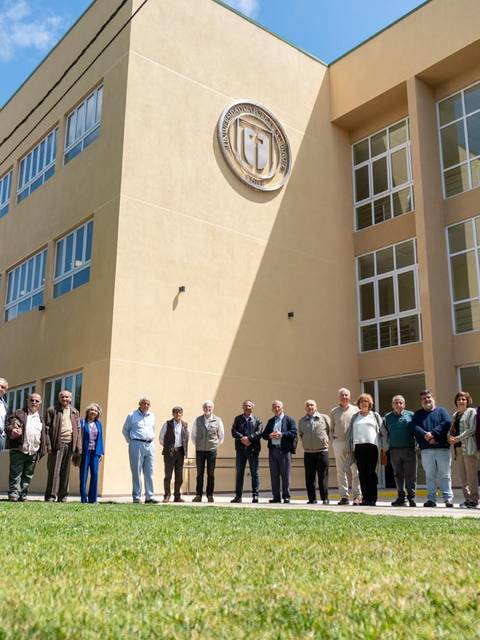 Un grupo de personas se encuentra alineado frente a un edificio con un gran logo institucional.