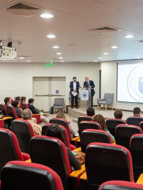 Una sala de conferencias con un presentador y un moderador frente a una audiencia sentada.