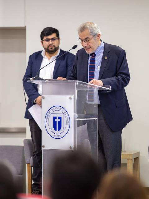 Un hombre mayor habla en un podio mientras otro hombre lo observa desde atrás.