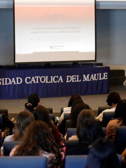 Una conferencia en la Universidad Católica del Maule con un ponente y una moderadora frente a un auditorio de estudiantes.