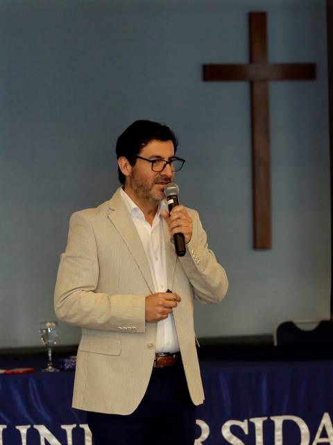 Un hombre con chaqueta clara presenta en un evento académico frente a un público.