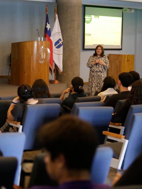 Una presentadora se encuentra hablando frente a un grupo de personas en un auditorio.