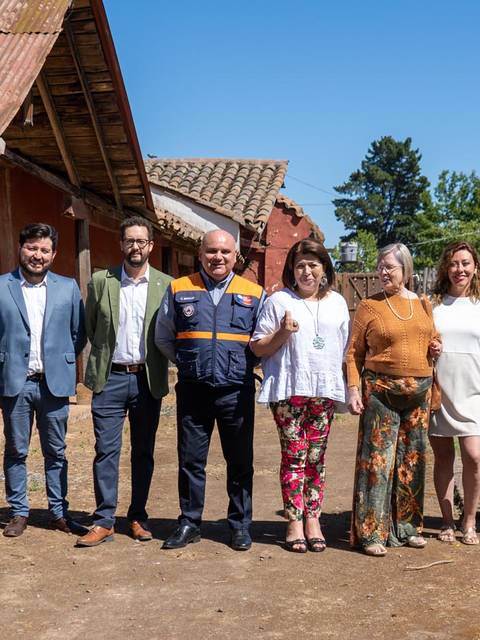Un grupo de personas posando frente a una casa rural con banderas de organizaciones en un día soleado.