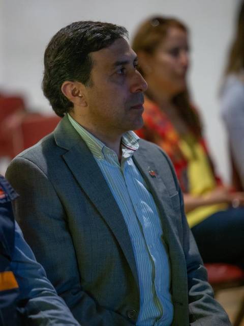 Un hombre sentado en un evento, escuchando atentamente mientras varias personas están presentes en el fondo.