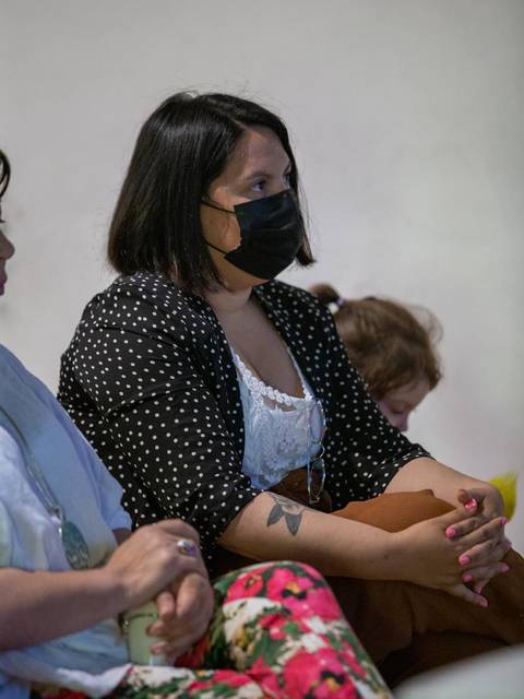 Una mujer sentada con una blusa blanca y chaqueta de lunares mientras lleva una mascarilla negra, en un ambiente tranquilo.