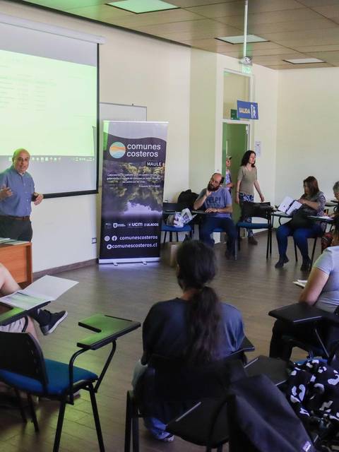 Un grupo de personas asiste a una presentación en un aula con un proyector y material visual sobre temas costeros.