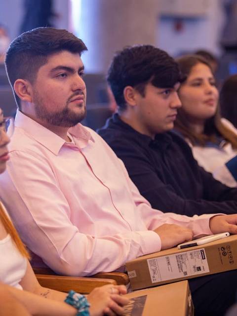 Un grupo de jóvenes está sentado en un auditorio, escuchando atentamente mientras sostienen cajas en sus manos.