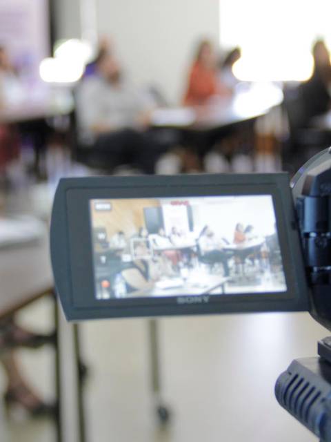 Una cámara graba una reunión en un aula con personas sentadas al fondo.
