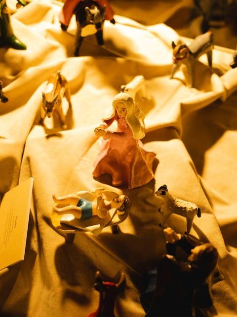 Una escena con figuras de plástico dispuestas sobre una tela iluminada, donde destaca una figura en vestido rosa en el centro.