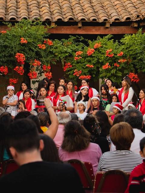 Un grupo de niños y niñas cantan en un evento navideño al aire libre rodeados de flores.