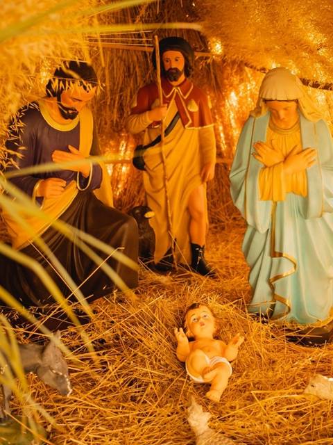 La imagen muestra una representación del nacimiento de Jesús con figuras de María, José y los Reyes Magos en un entorno de paja iluminado.