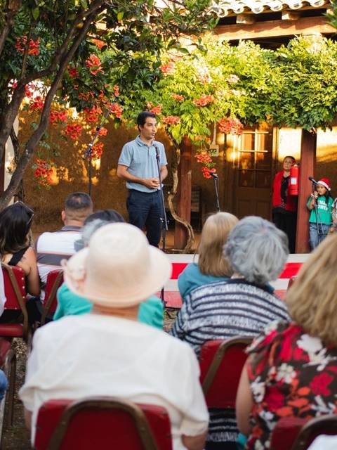 Un hombre está hablando frente a un público en un evento al aire libre rodeado de árboles y flores.