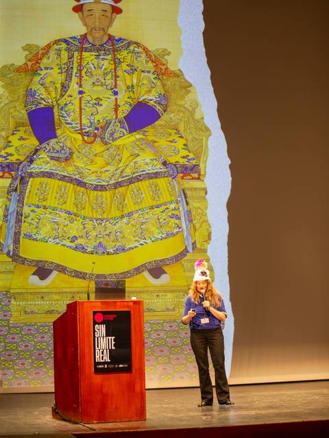 Una mujer habla en un escenario mientras una imagen de un emperador chino aparece de fondo.