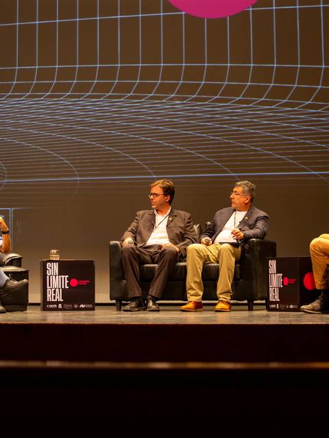 Una charla en un escenario con cuatro panelistas sentados en sillas de cuero con un fondo gráfico.