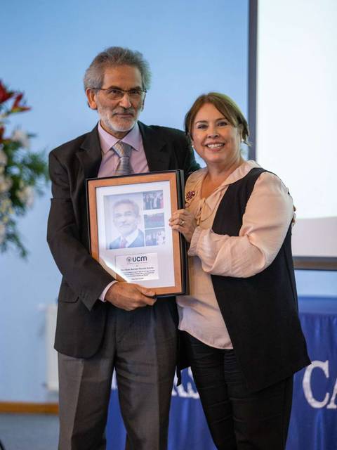 En la imagen, dos personas sostienen un marco con una fotografía en un evento formal.