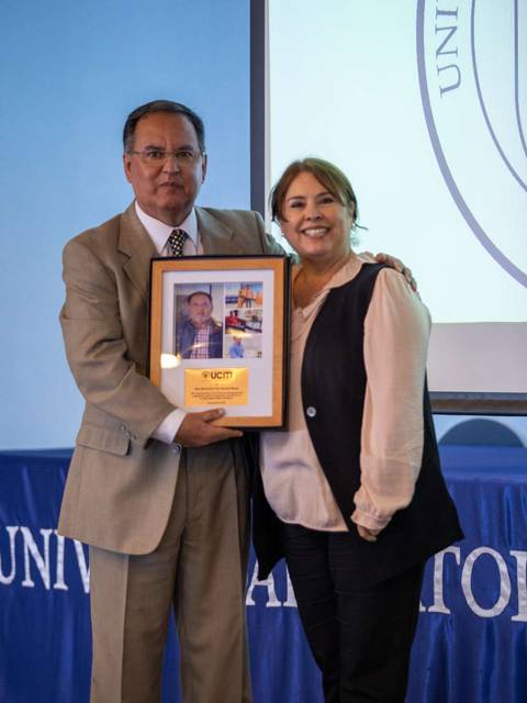 Dos personas están posando juntas, sosteniendo un marco con una foto en un entorno académico.