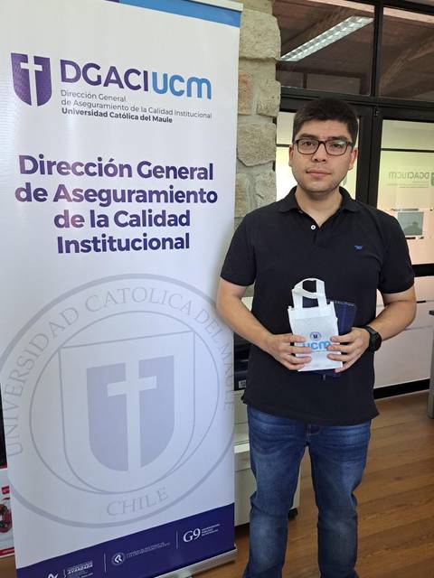 Una persona sosteniendo un folleto frente a un banner institucional en un entorno de oficina.