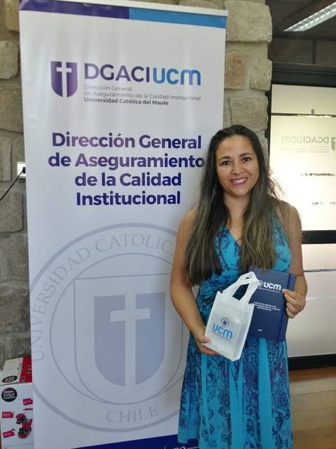 Una mujer sonriente sostiene una bolsa y un cuaderno frente a un banner institucional en un entorno de oficina.