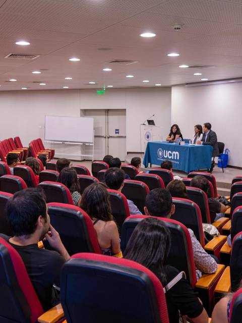 Una sala de conferencias con asientos rojos ocupados por un público que escucha a un panel de tres personas en el escenario.