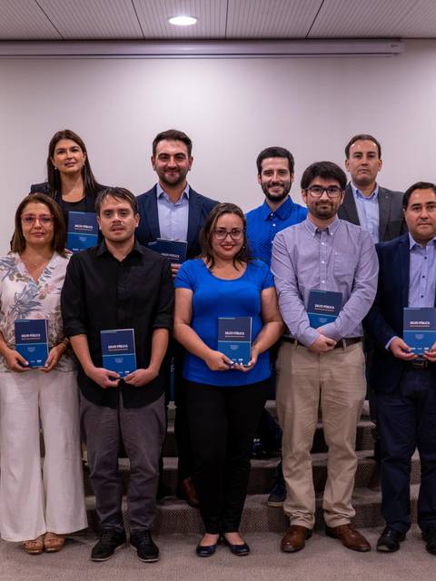 Un grupo de personas sonrientes sosteniendo libros en un evento formal en un salón con banderas.