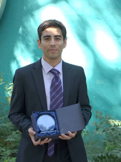 Un hombre joven vestido con traje sostiene un objeto en una caja mientras sonríe frente a un fondo verde.