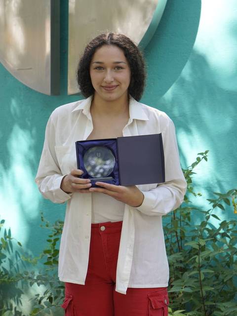 Una joven sonriente sostiene un objeto dentro de una caja mientras está frente a un fondo verde.